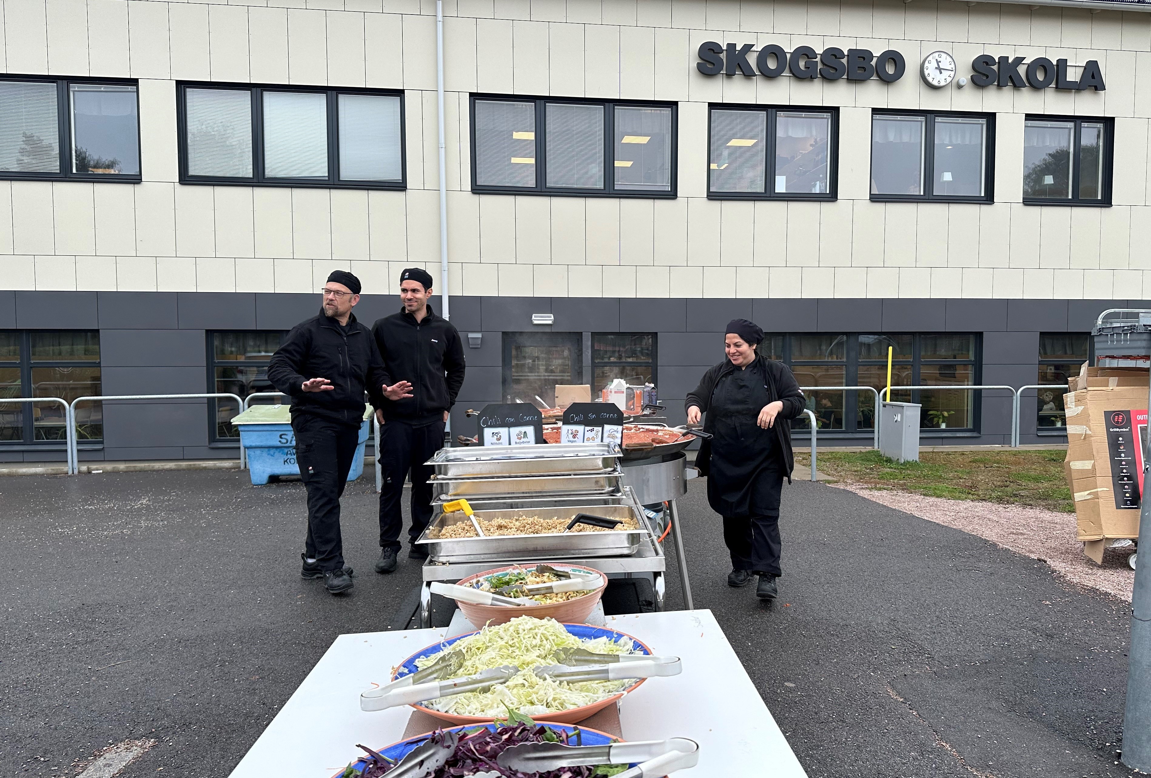 Skolpersonal står vid matlagningsstation utomhus