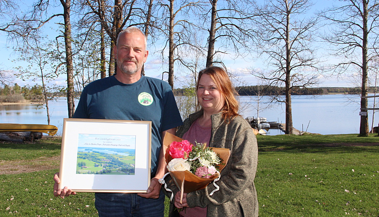 Två personer stående med en tavla och blommor med en sjö bakom sig.
